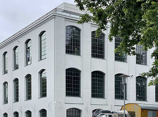 Trockenbau Kaufmann Alte Spinnerei Mehrstöckiges, weißes Gebäude mit großen, rechteckigen Fenstern und Bäumen im Vordergrund.