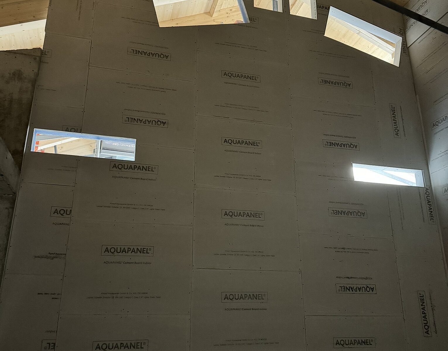 Innenaufnahme einer Baustelle mit einer Wand, die mit AQUAPANEL-Platten verkleidet ist, darunter mehrere schmale Fenster, durch die Tageslicht einfällt, und sichtbare Holzbalken der Dachkonstruktion.