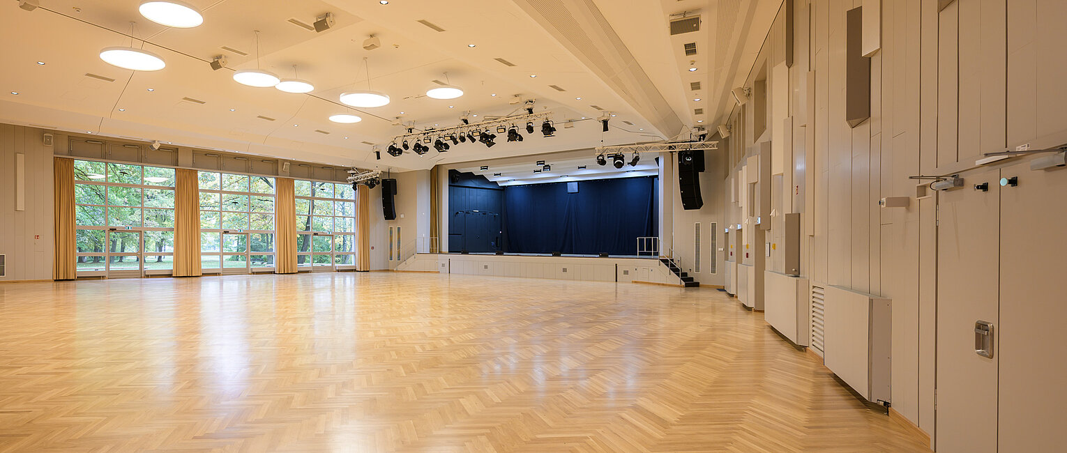 Großer leerer Saal mit heller Holzboden, Bühne mit blauen Vorhängen, großen Fenstern und modernen Deckenleuchten.