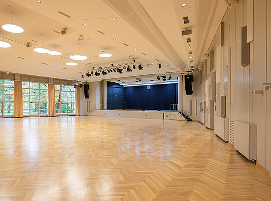 Trockenbau Kaufmann Stadthalle Weilheim Großer leerer Saal mit heller Holzboden, Bühne mit blauen Vorhängen, großen Fenstern und modernen Deckenleuchten.
