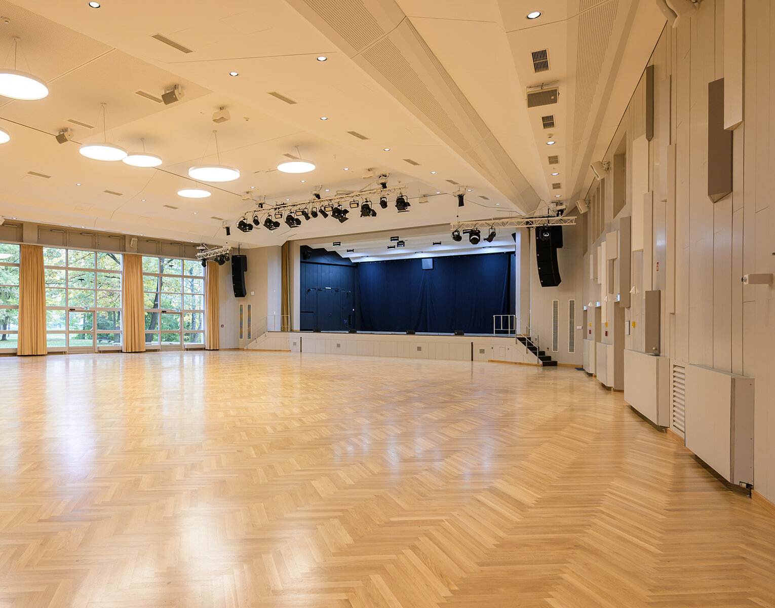 Großer leerer Saal mit heller Holzboden, Bühne mit blauen Vorhängen, großen Fenstern und modernen Deckenleuchten.