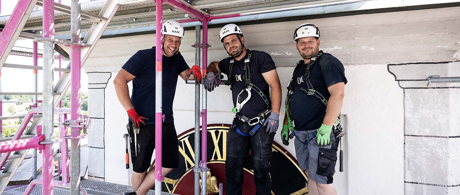 Drei Bauarbeiter mit Schutzhelmen und Sicherheitsausrüstung stehen auf einem Gerüst vor einem großen Uhrenzifferblatt und lächeln in die Kamera.