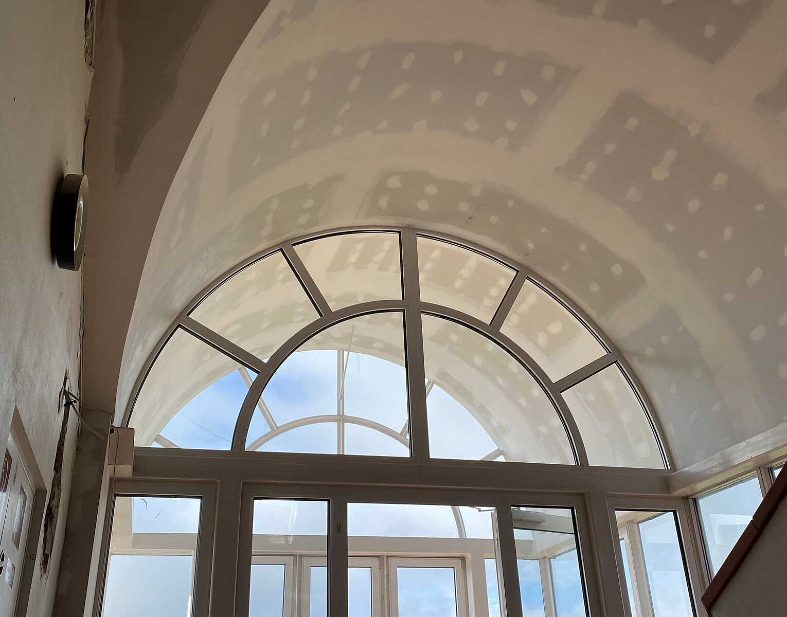 Blick auf große Fensterfront mit Rundbogen im oberen Bereich, viel Tageslicht fällt in einen Innenraum mit glattem, gewölbtem und teils unfertigem Dach. Draußen sind Bäume und Himmel sichtbar.