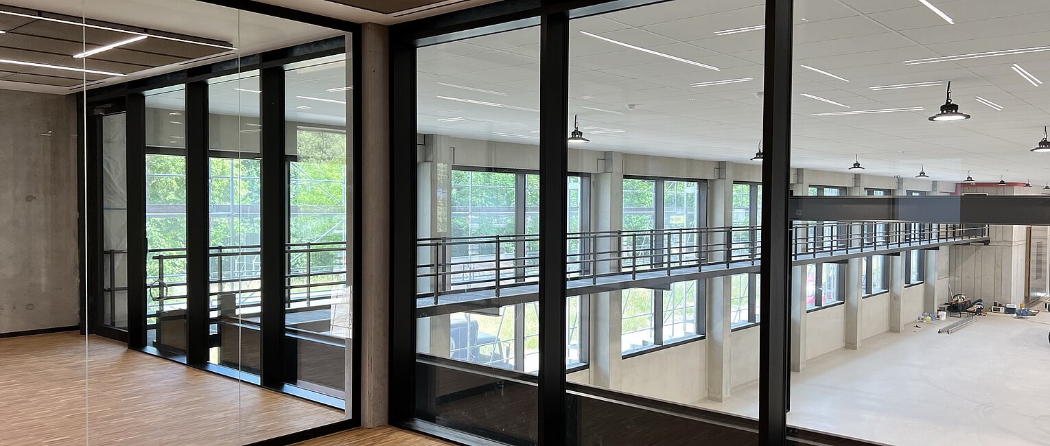Blick in einen modernen Innenraum mit großen Glaswänden, Holzboden und einem hellen, offenen Bereich mit hoher Decke und zahlreichen Fenstern im Hintergrund.