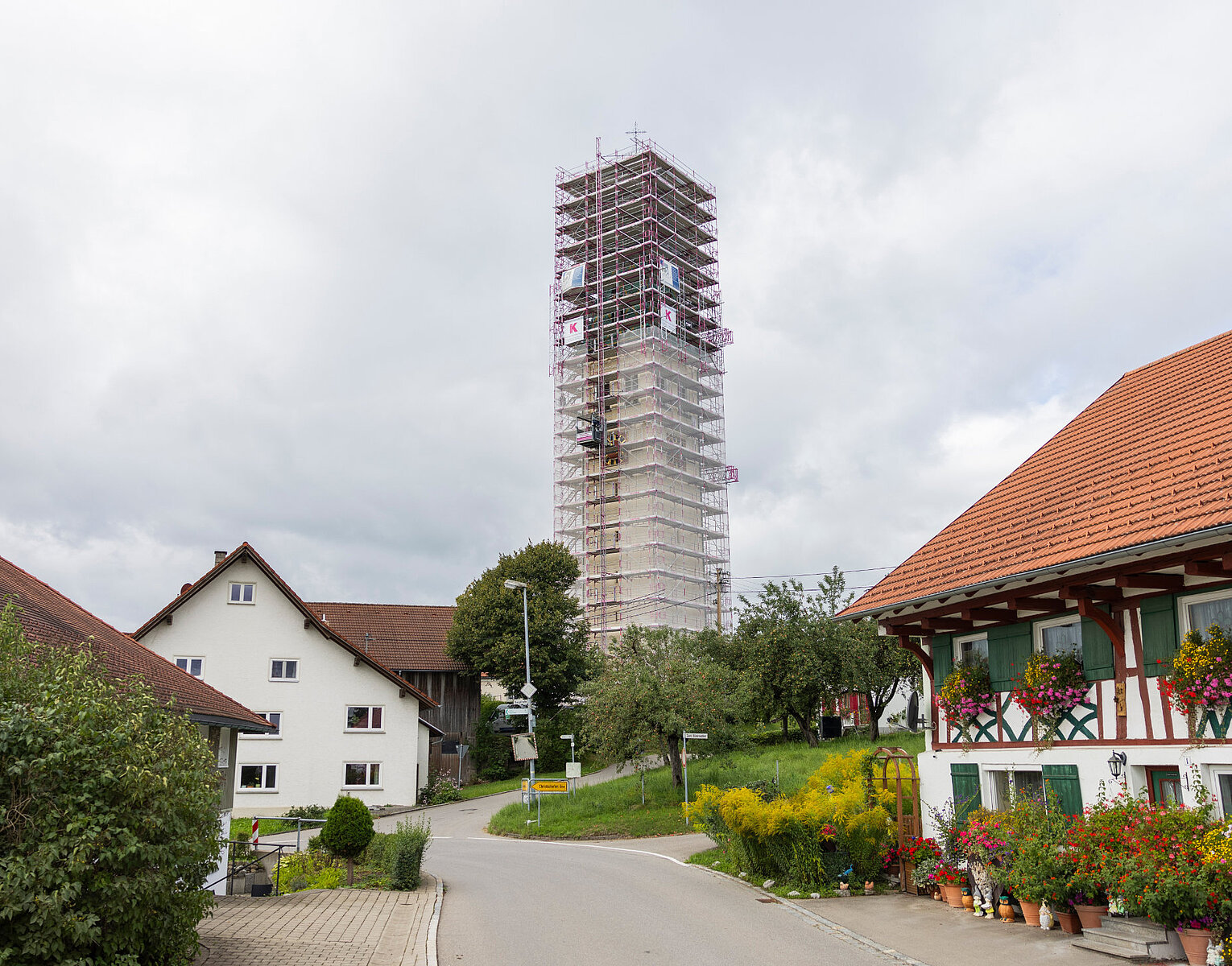Dorfgasse mit Fachwerkhaus und blühendem Garten, im Hintergrund ein hoher Kirchturm, der von einem Gerüst umgeben ist, unter bewölktem Himmel.
