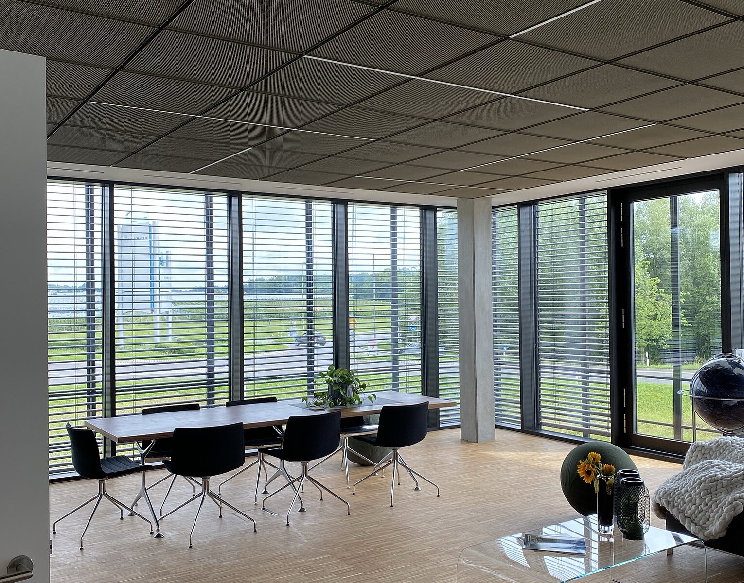 Moderner Besprechungsraum mit großem Tisch, mehreren schwarzen Stühlen, großen Fenstern mit Jalousien und Blick auf grüne Außenanlagen. Im Vordergrund ein Sofa mit Decke und ein kleiner Glastisch mit Blumen.