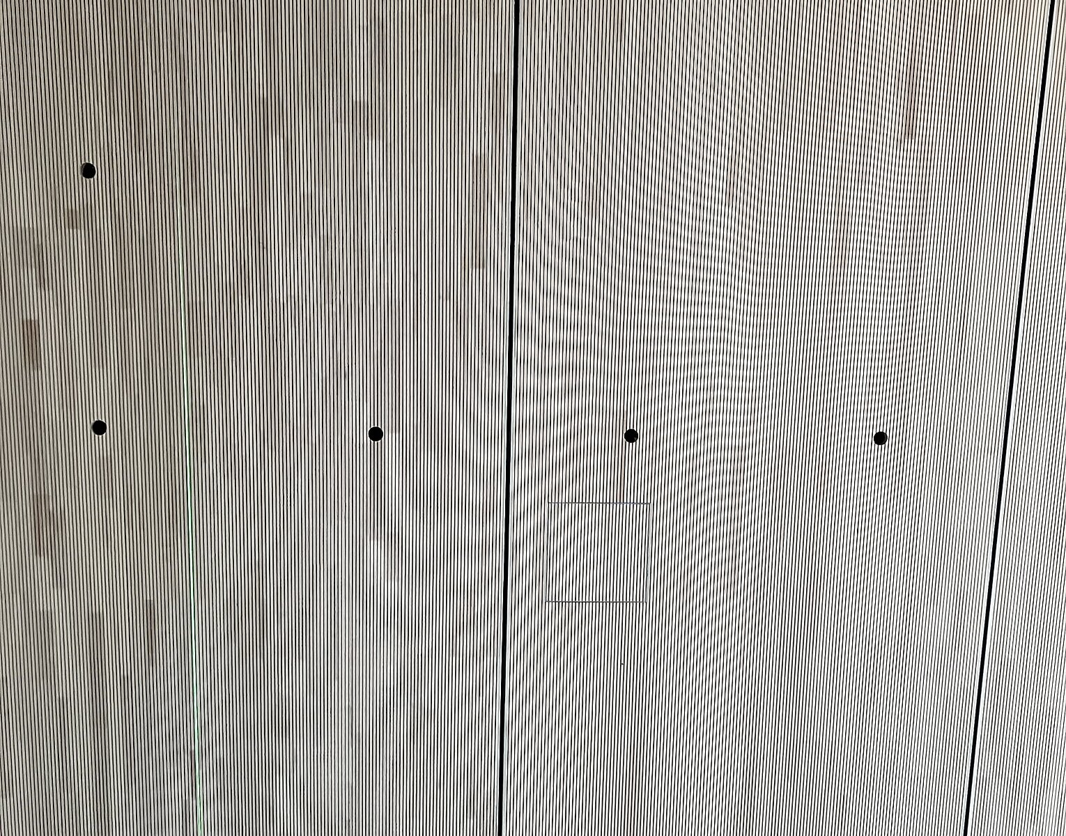 Wand aus hellen Holzpaneelen mit schwarzen Löchern und dünnen vertikalen Spalten, davor ein kleiner Teil eines Trolleys mit schwarzem Griff.