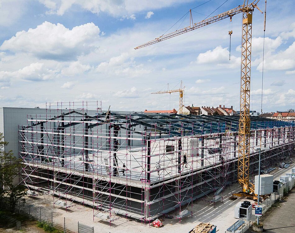 Großes Baustellengelände mit einem Rohbau, umgeben von Gerüsten und zwei Baukränen, unter einem bewölkten Himmel.