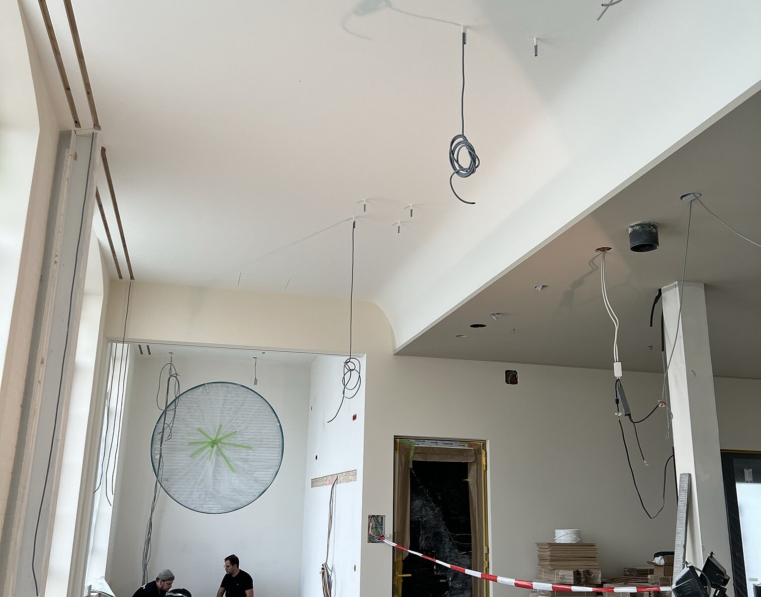 Innenraum einer Baustelle mit hohen Fenstern, nackten Kabeln an der Decke, Bauarbeiter in der Ecke und Baumaterialien auf dem Boden verteilt.