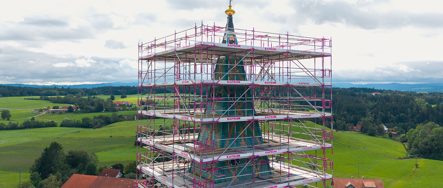 Kirchturm eines Gebäudes, umgeben von einem Gerüst, in einer ländlichen Gegend mit grünen Feldern und Bauernhäusern im Hintergrund.