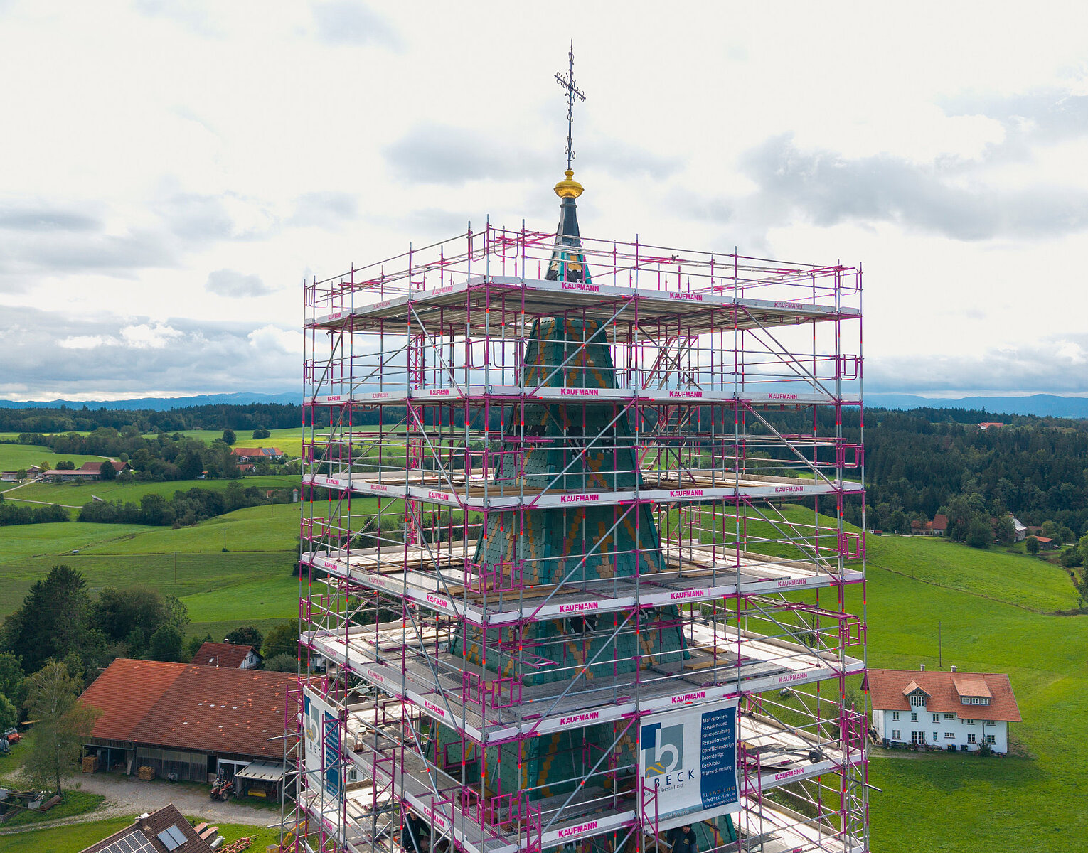 Kirchturm eines Gebäudes, umgeben von einem Gerüst, in einer ländlichen Gegend mit grünen Feldern und Bauernhäusern im Hintergrund.