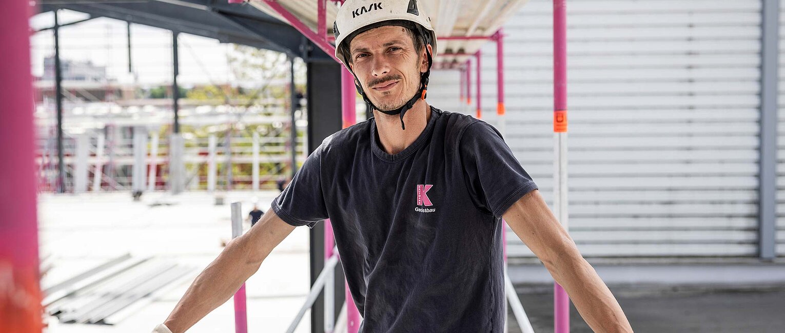 Bauarbeiter mit Helm und Handschuhen steht auf einer Baustelle zwischen Gerüststangen und blickt in die Kamera.
