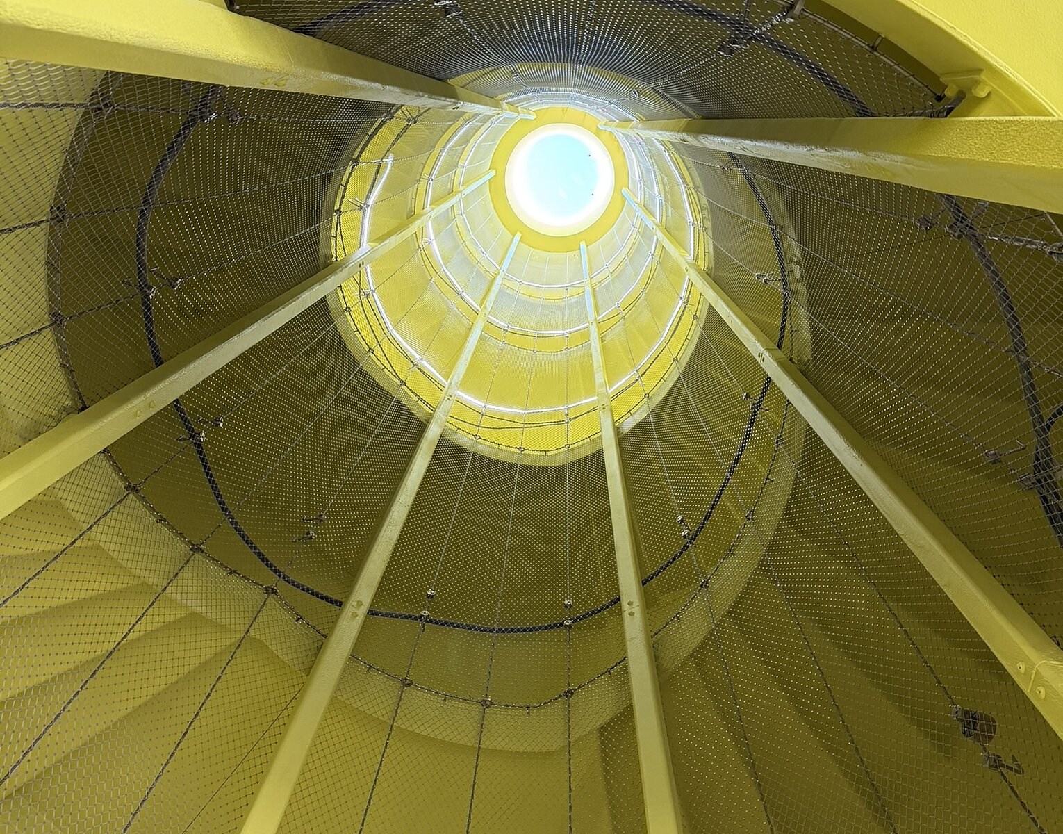 Blick nach oben auf eine gelbe, spiralförmige Wendeltreppe mit Geländern und Netzstrukturen, die sich zu einem runden, hell erleuchteten Öffnungspunkt am Himmel verjüngt.