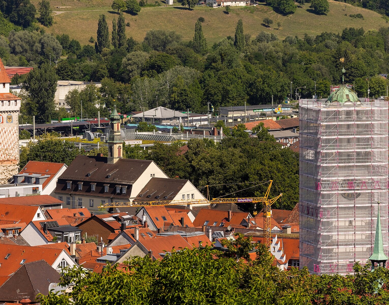 Stadtansicht mit roten Dächern, einem historischen Turm links und einem Turm rechts, der von einem Gerüst umgeben ist, im Hintergrund grüne bewaldete Hügel.