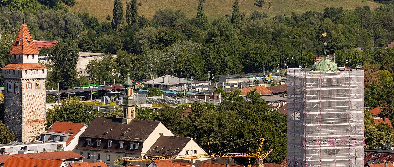 Stadtansicht mit roten Dächern, einem historischen Turm links und einem Turm rechts, der von einem Gerüst umgeben ist, im Hintergrund grüne bewaldete Hügel.