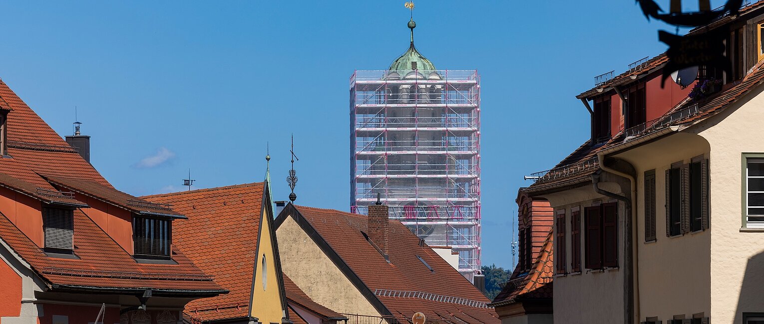 Blick auf eine historische Altstadt mit farbigen Häusern und einem Turm im Hintergrund, der von Gerüsten für Renovierungsarbeiten umgeben ist.