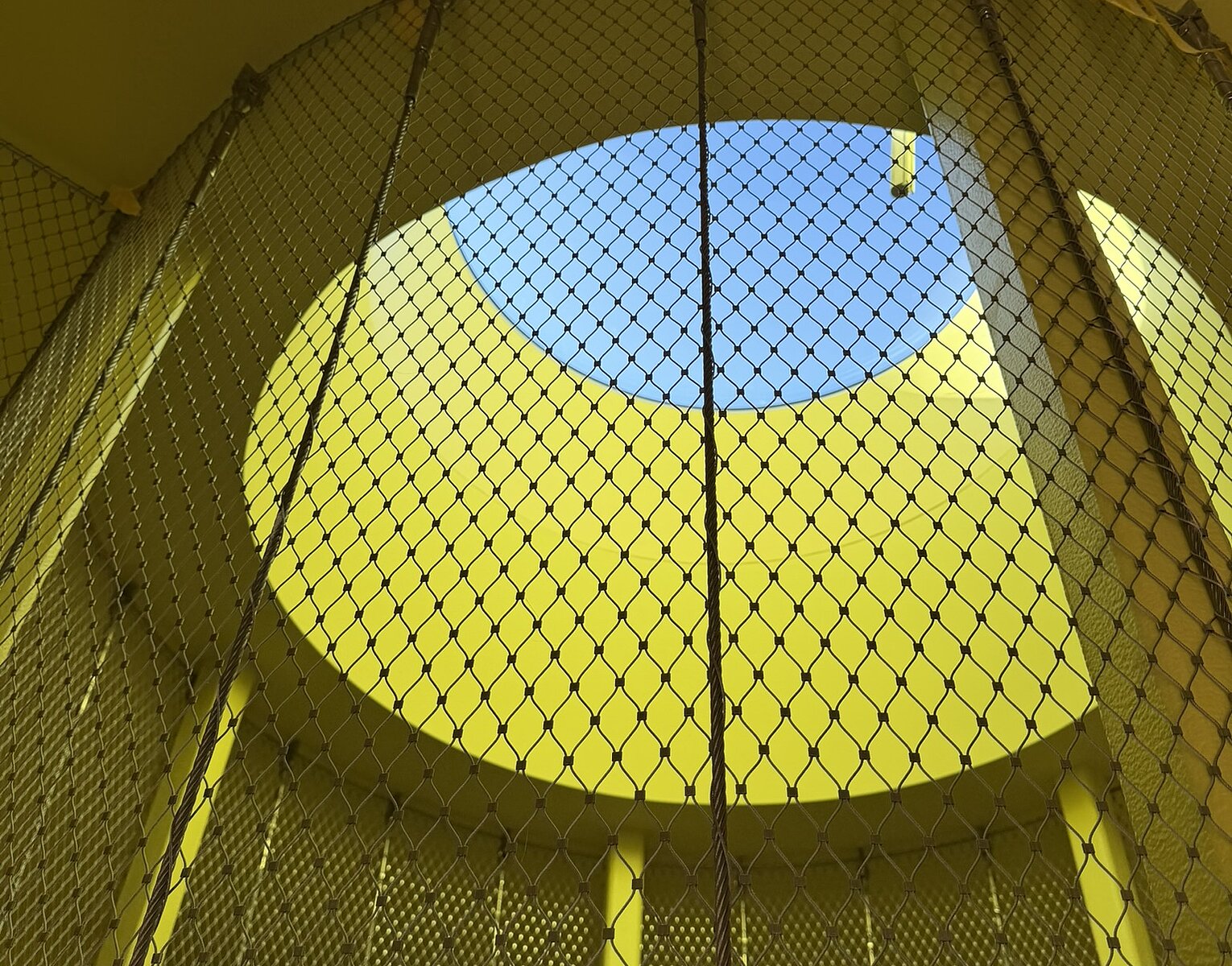 Blick nach oben durch ein rundes, gelbes Treppenhaus mit einem Gitterzaun und offenem Himmel in der Mitte.
