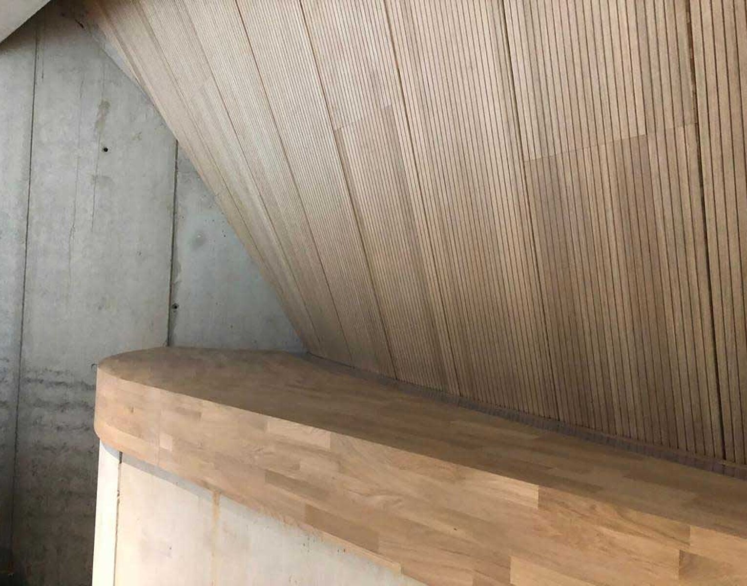 Moderner Innenraum mit einer Holzbank und einer schrägen Wandverkleidung aus dünnen Holzlatten neben einer Betonwand.