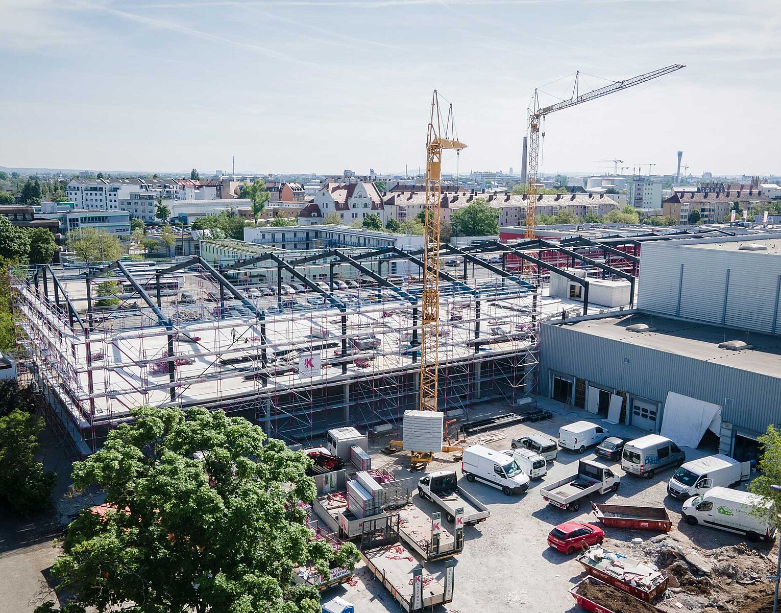 Bau einer großen Gebäudestruktur mit Stahlrahmen, mehreren Kränen und Baumaterialien auf einer Baustelle in einer urbanen Umgebung bei Tageslicht.