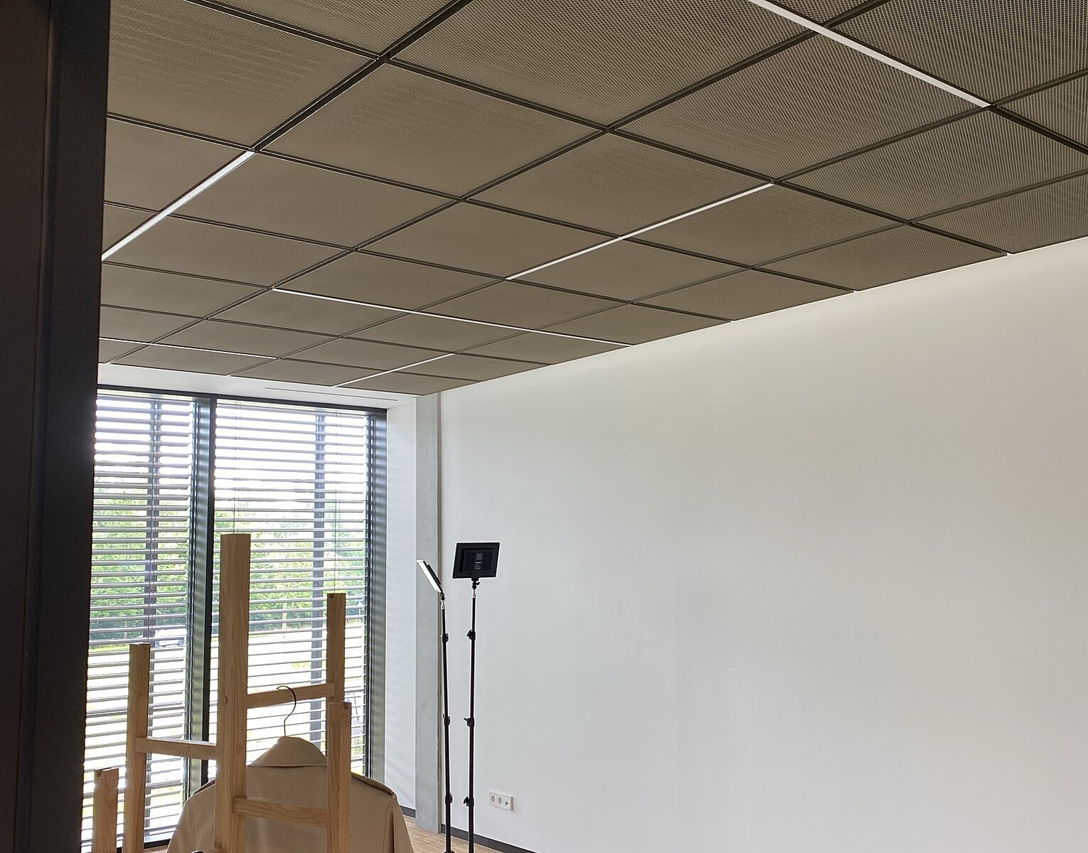 Leerer Raum mit weißer Wand, Holzkleiderständer und einem beleuchteten Stativ in der Ecke vor einem großen Fenster mit Jalousien.