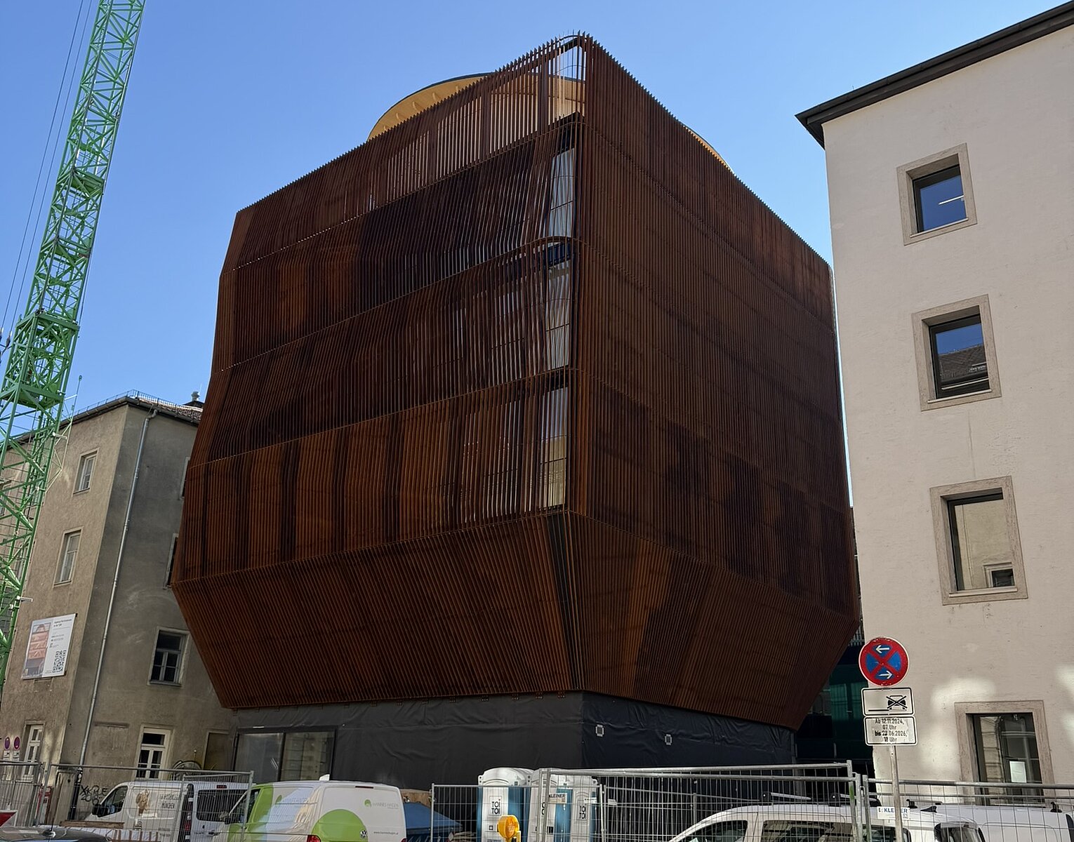 Modernes braun verkleidetes Gebäude zwischen älteren Gebäuden, Baustelle mit Bauzaun und geparkten Fahrzeugen im Vordergrund, blauer Himmel.