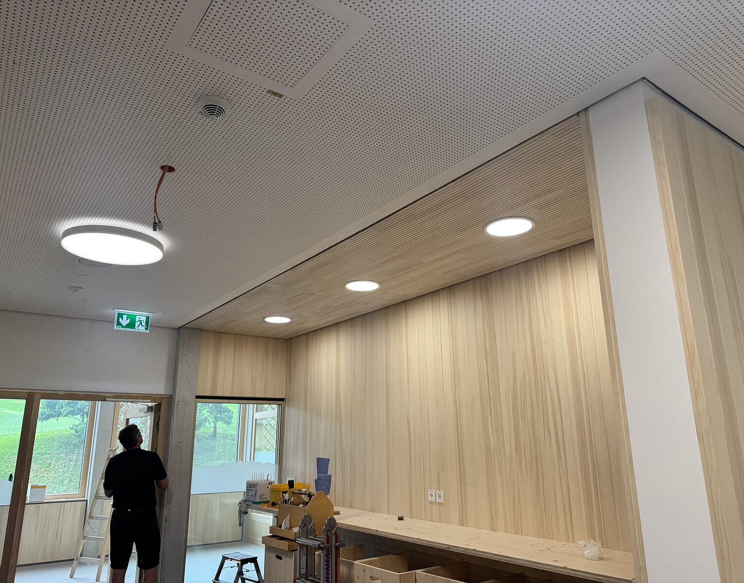 Innenraum eines modernen Raums mit Holzvertäfelungen, Deckenbeleuchtung und einer Person in dunkler Kleidung, die an der Tür steht.