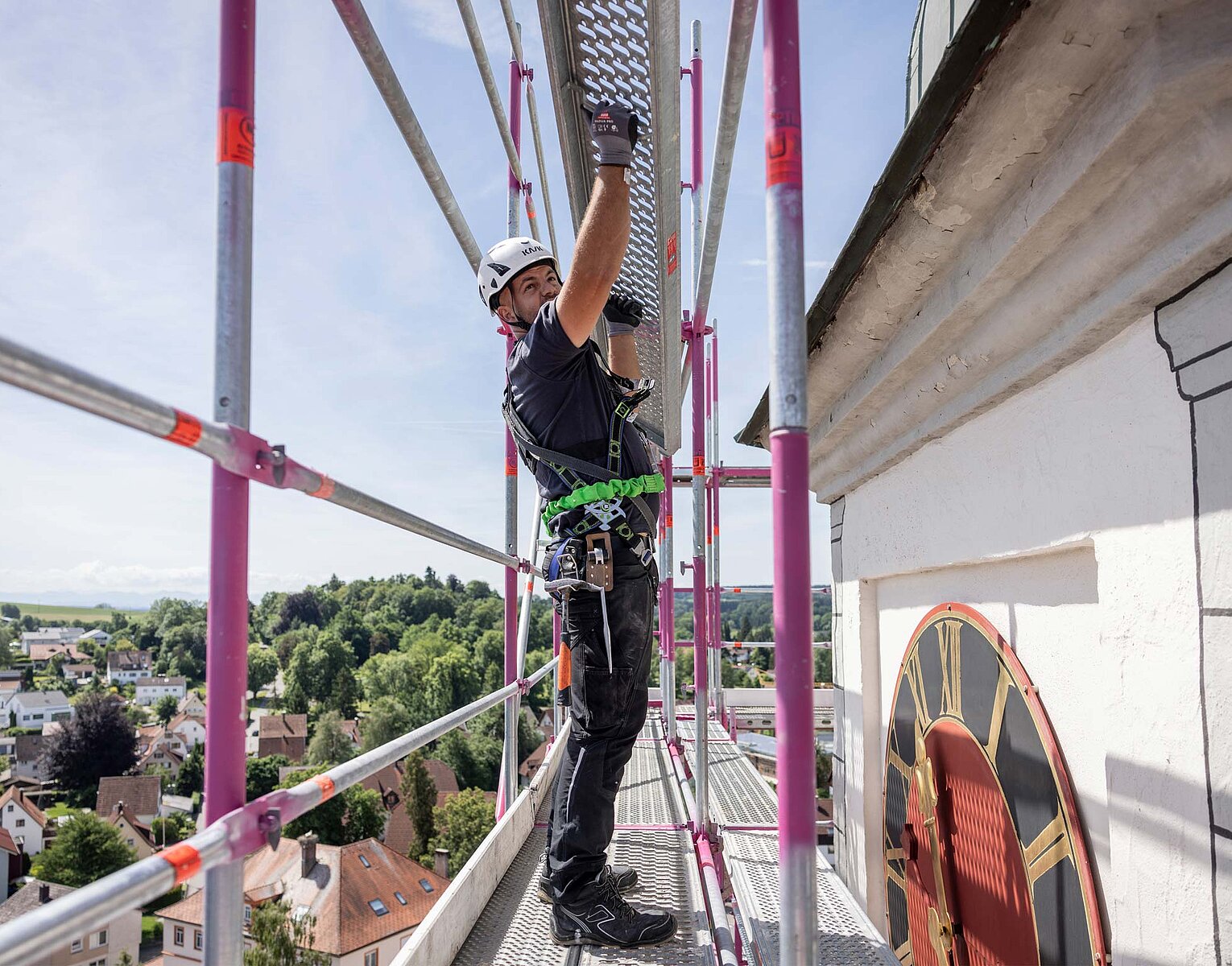 Ein Handwerker mit Helm und Sicherheitsgurt arbeitet auf einem Gerüst an der Außenwand eines historischen Gebäudes mit einer großen Uhr. Im Hintergrund ist eine Stadt und Landschaft sichtbar.