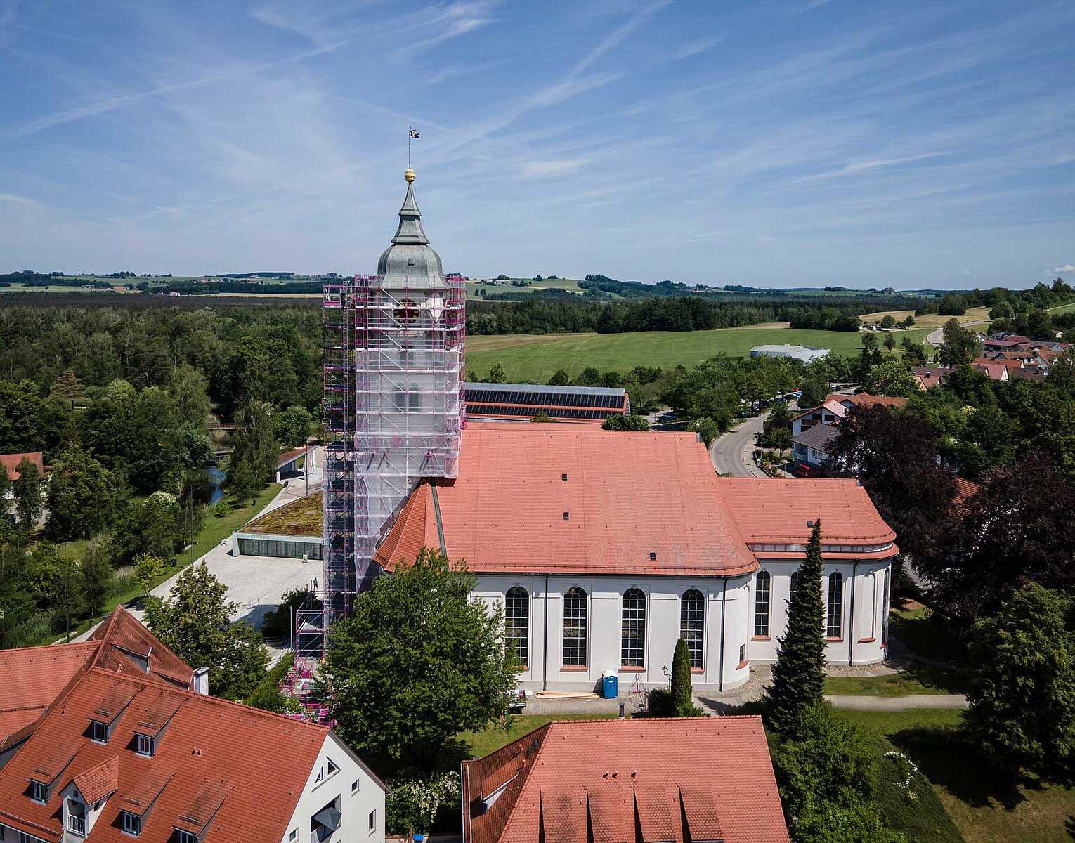 Blick auf eine Kirche mit rotem Dach, deren Turm von Gerüsten umgeben ist, im ländlichen Gebiet mit umgebenden Häusern, Bäumen und Feldern unter blauem Himmel.