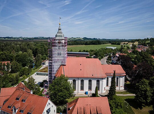 Blick auf eine Kirche mit rotem Dach, deren Turm von Gerüsten umgeben ist, im ländlichen Gebiet mit umgebenden Häusern, Bäumen und Feldern unter blauem Himmel.