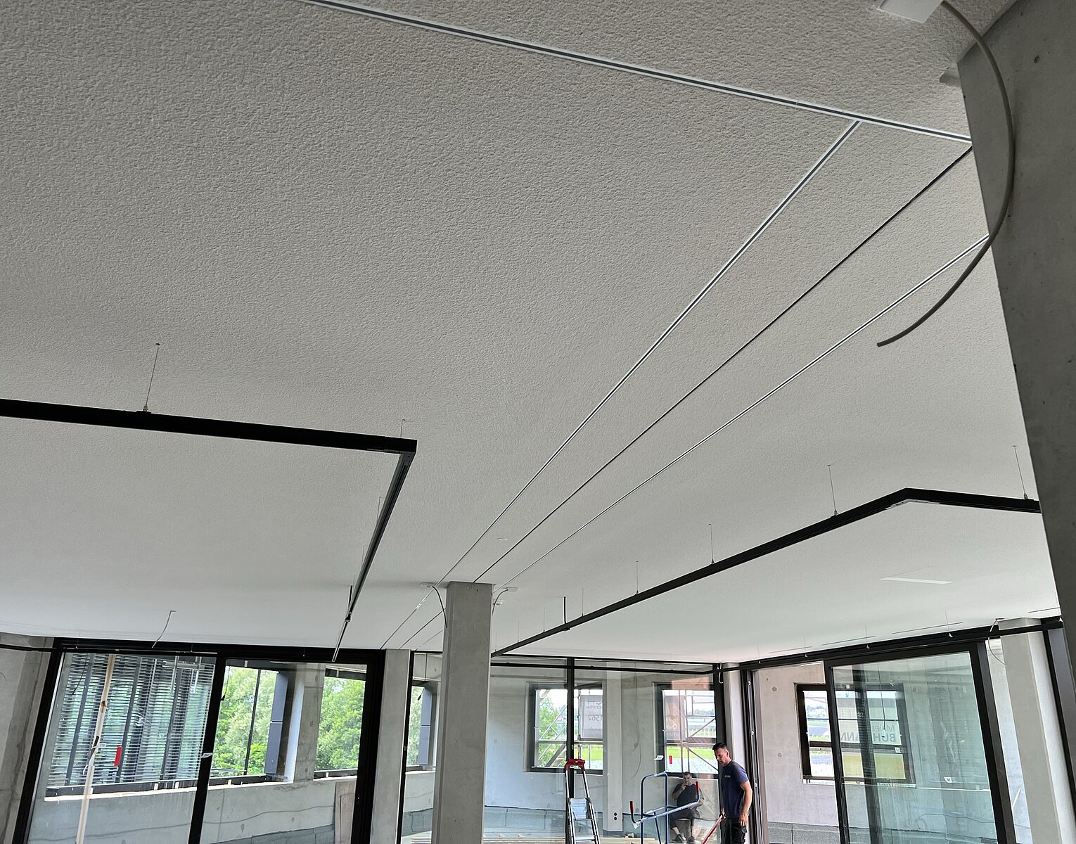 Innenraum eines modernen Gebäudes im Rohbau mit offenem Grundriss, großen Fenstern und sichtbaren Betonstützen, eine Person arbeitet an einer Leiter nahe einem Fenster.