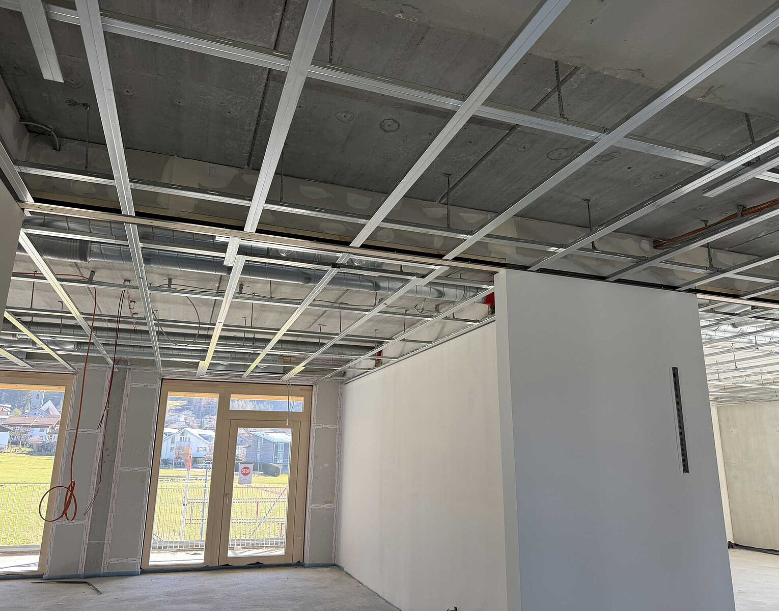Innenraum eines Gebäudes im Rohbau mit sichtbarer Deckenunterkonstruktion aus Metall und verputzten Wänden, große Fenster mit Blick ins Freie, Boden und Decke noch nicht fertiggestellt.