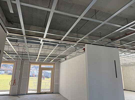Trockenbau Kaufmann KITA Burgberg Innenraum eines Gebäudes im Rohbau mit sichtbarer Deckenunterkonstruktion aus Metall und verputzten Wänden, große Fenster mit Blick ins Freie, Boden und Decke noch nicht fertiggestellt.