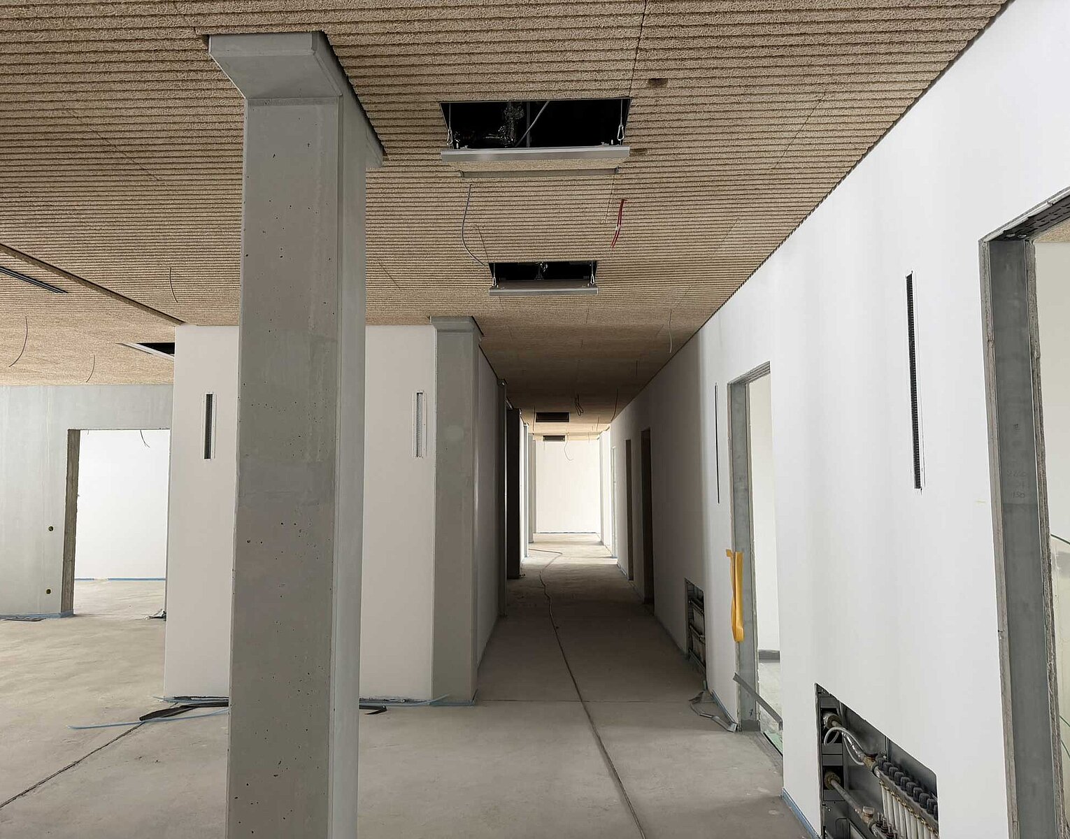 Blick in einen breiten Flur eines unvollendeten Gebäudes mit weißen Wänden und offenen Deckenplatten, sichtbaren Kabeln und Rohren, ohne Bodenbelag und mit einem Betonsäule in der Mitte.