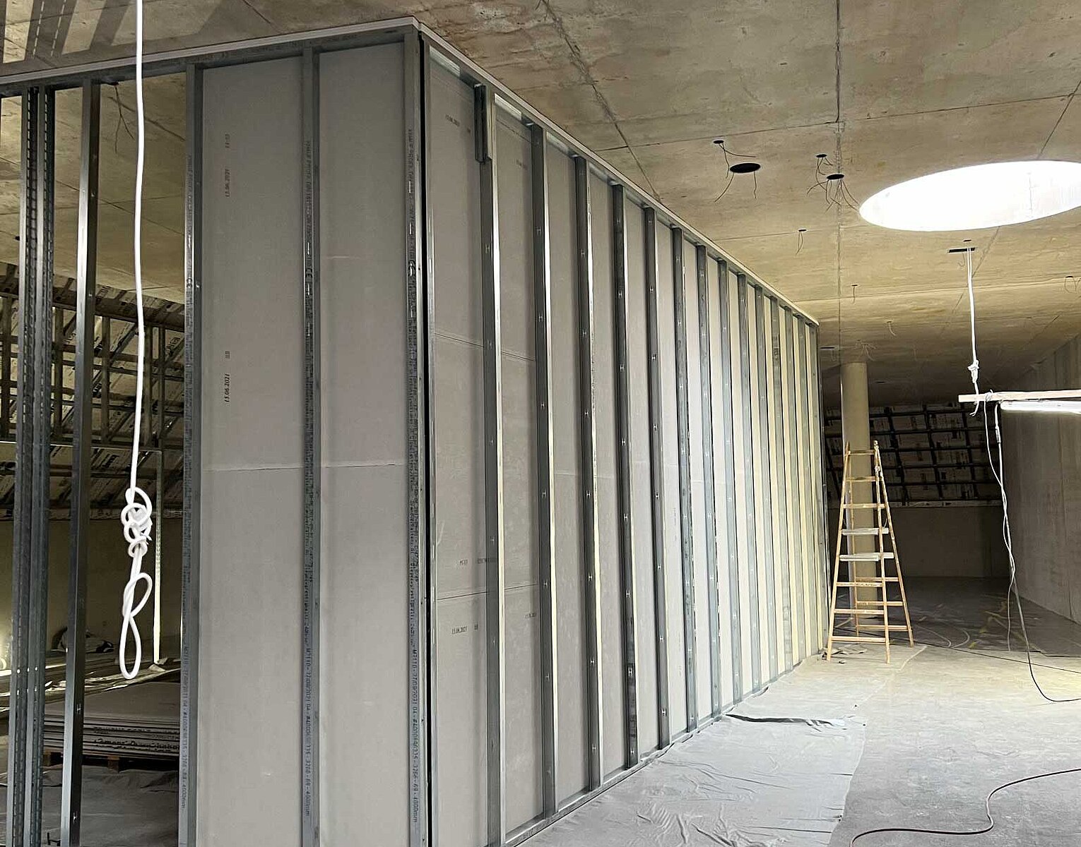 Innenansicht einer Baustelle mit aufgehängten Kabeln, einer Treppe im Hintergrund und einer halb fertigen Trennwand aus Metallständern und Gipskartonplatten.