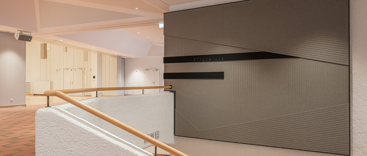Innenansicht eines modernen Gebäudes mit einer Treppe, Holzgeländer und einer großen, abstrakten Wandkunst aus schwarzen Linien auf grauem Hintergrund.