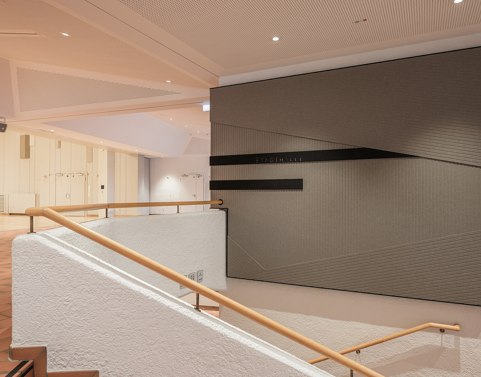 Innenansicht eines modernen Gebäudes mit einer Treppe, Holzgeländer und einer großen, abstrakten Wandkunst aus schwarzen Linien auf grauem Hintergrund.