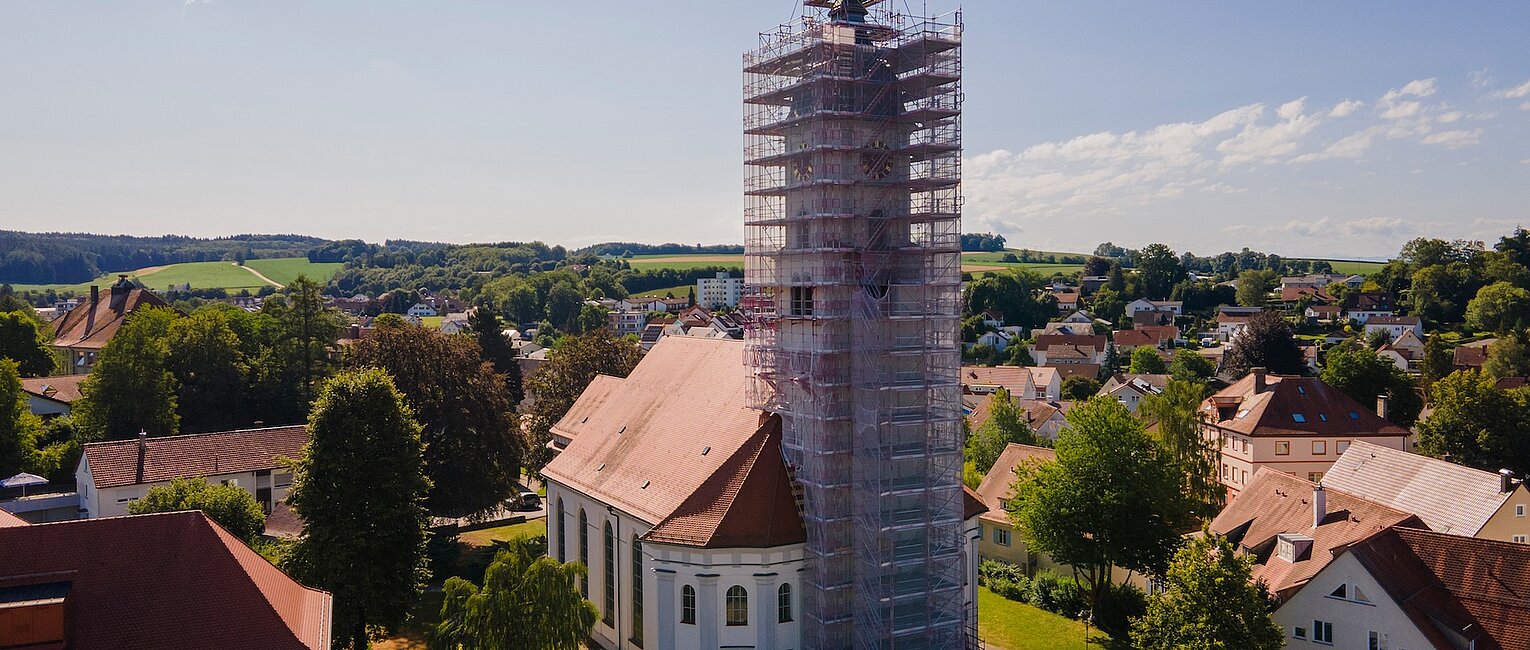 Kirche mit einem Turm, der von einem Gerüst für Renovierungsarbeiten umgeben ist, umgeben von Bäumen und Häusern in einer ländlichen Stadtlandschaft unter klarem Himmel.