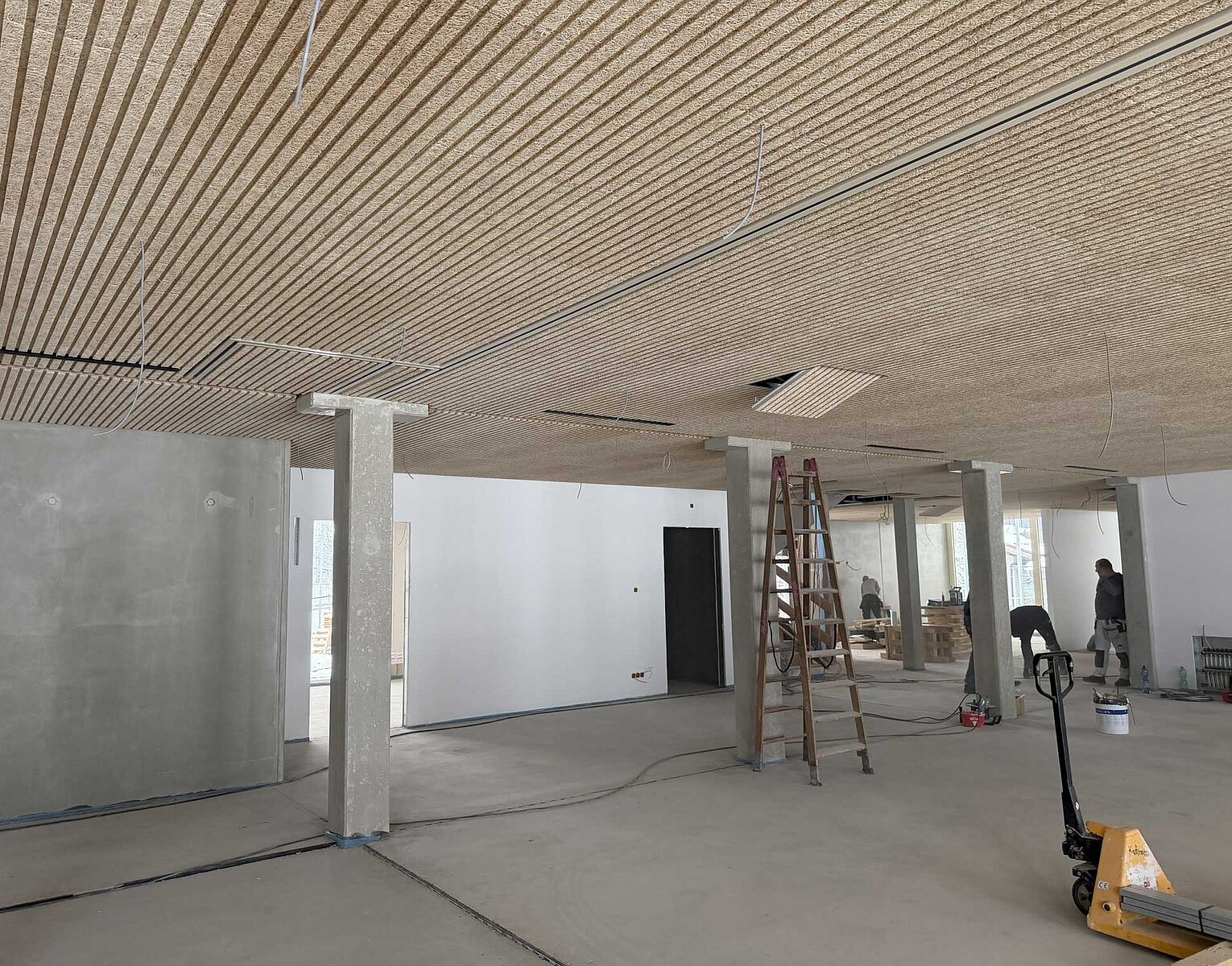 Großer Raum in einem Gebäude im Rohbauzustand mit sichtbaren Deckenplatten, Betonstützen, einer Leiter und Bauwerkzeugen auf dem Boden.