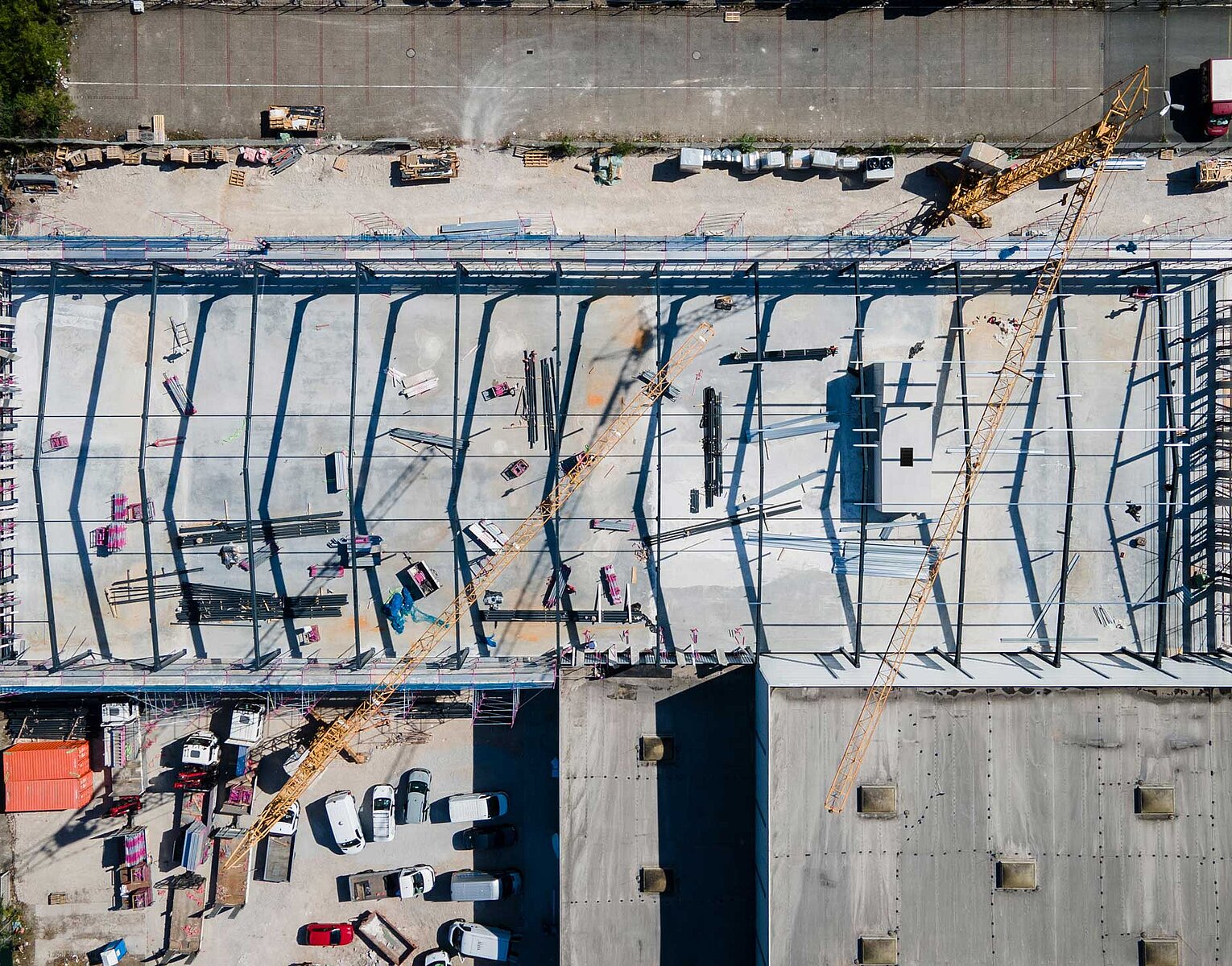 Luftaufnahme einer Baustelle mit Stahlrahmenstruktur eines großen Gebäudes und mehreren Kränen auf einem betonierten Fundament. Fahrzeuge und Baumaterialien sind rund um die Baustelle verteilt.