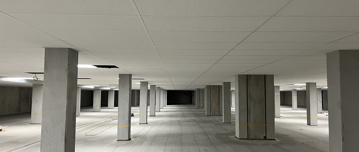 Großer leerer Tiefgaragenraum mit Betonsäulen, glattem Boden und Decke, beleuchtet durch Deckenlampen.