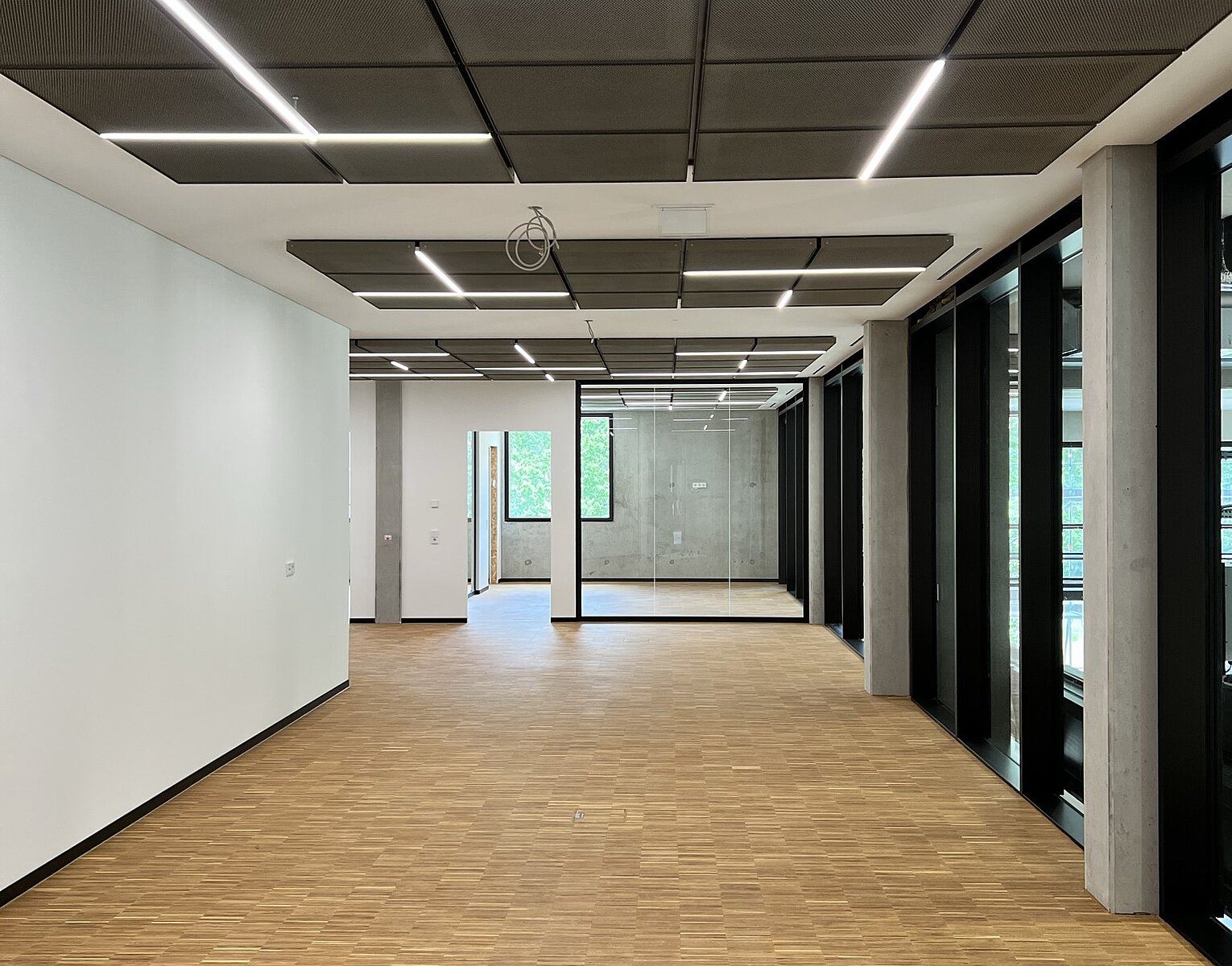 Leerer moderner Büroraum mit Holzboden, weißen und gläsernen Wänden sowie eingebauten Deckenleuchten in einem geometrischen Muster.