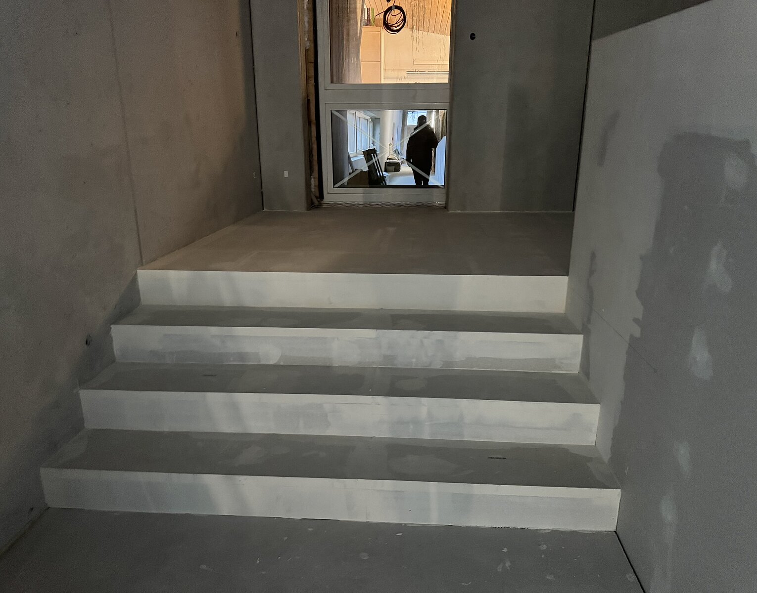Leere Treppe aus Beton in einem unfertigen Innenraum mit Sicht auf einen Flur und eine Person durch eine Glasscheibe.