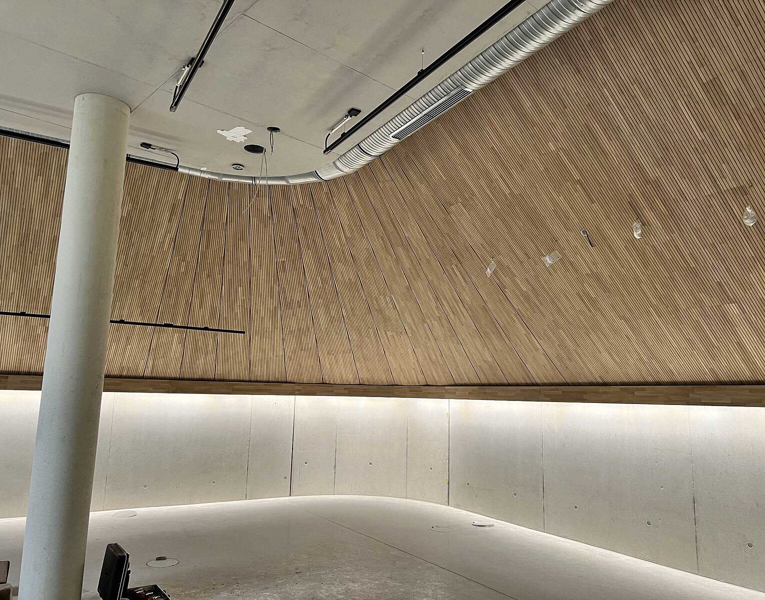 Innenraum mit geschwungener Holzvertäfelung an der Wand, weißem Betonboden und sichtbaren Rohrleitungen an der Decke.