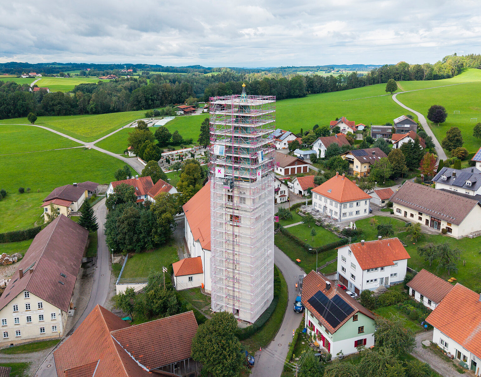 Luftaufnahme eines kleinen Dorfes mit einem hohen Turm in Gerüstbauweise inmitten von Häusern und grünen Wiesen unter bewölktem Himmel.