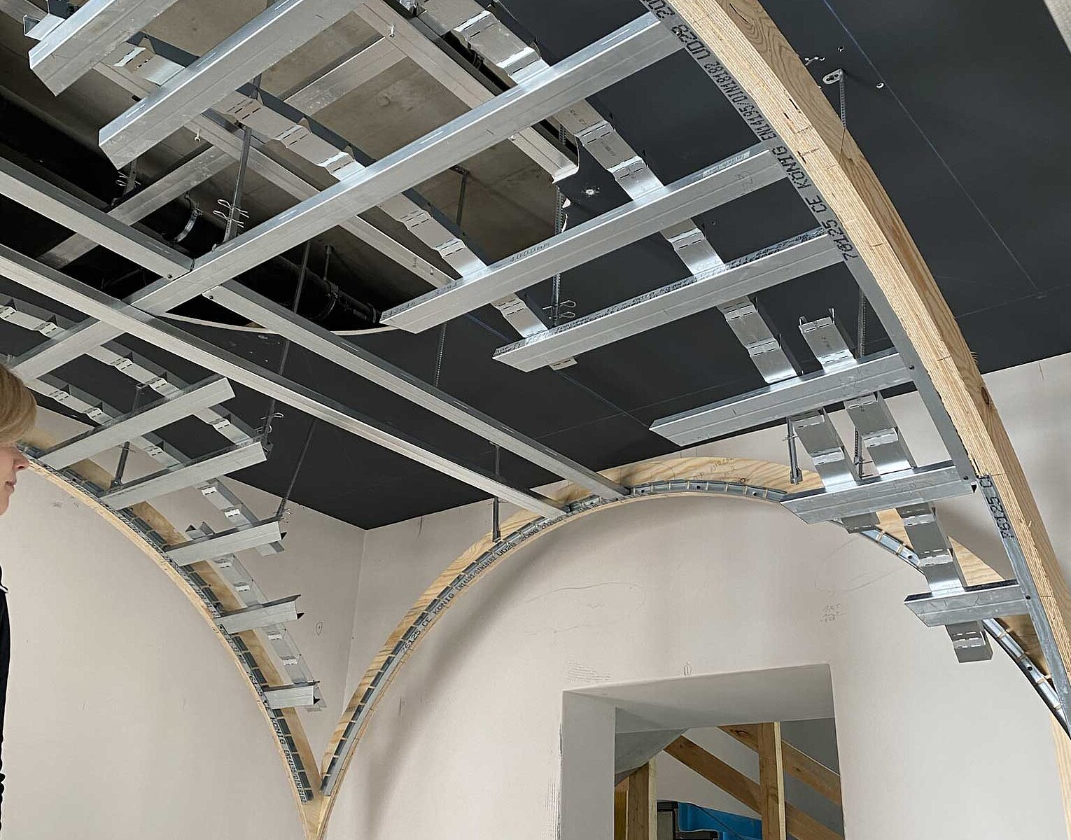 Innenansicht eines Raums mit sichtbarer Deckenstruktur aus Holz- und Metallrahmen, die für den Einbau einer abgehängten Decke vorbereitet wird. Im Hintergrund ist eine Treppe mit blauen Stufen teilweise sichtbar.