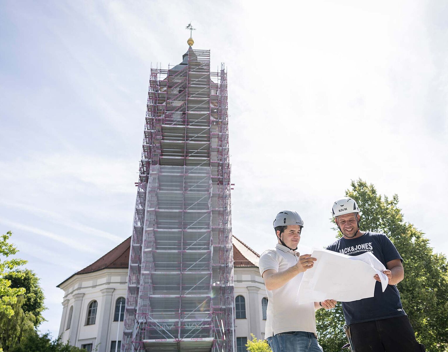 Zwei Bauarbeiter mit Helmen betrachten einen Bauplan vor einem hohen Kirchturm, der von Gerüsten umgeben ist.