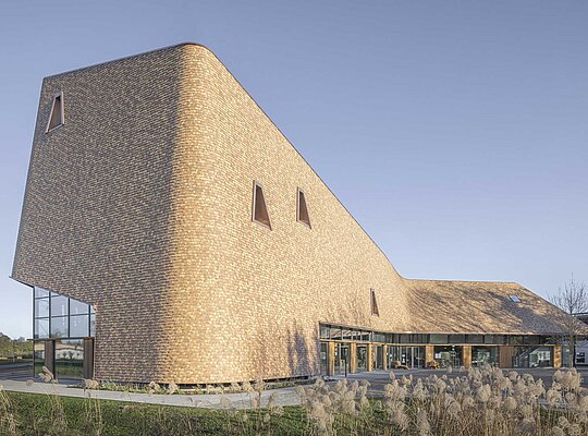 Trockenbau Kaufmann Rapunzel Besucher Zentrum Modernes Gebäude mit geschwungener Fassade aus Holzschindeln und kleinen dreieckigen Fenstern, umgeben von Gras und offenem Himmel.