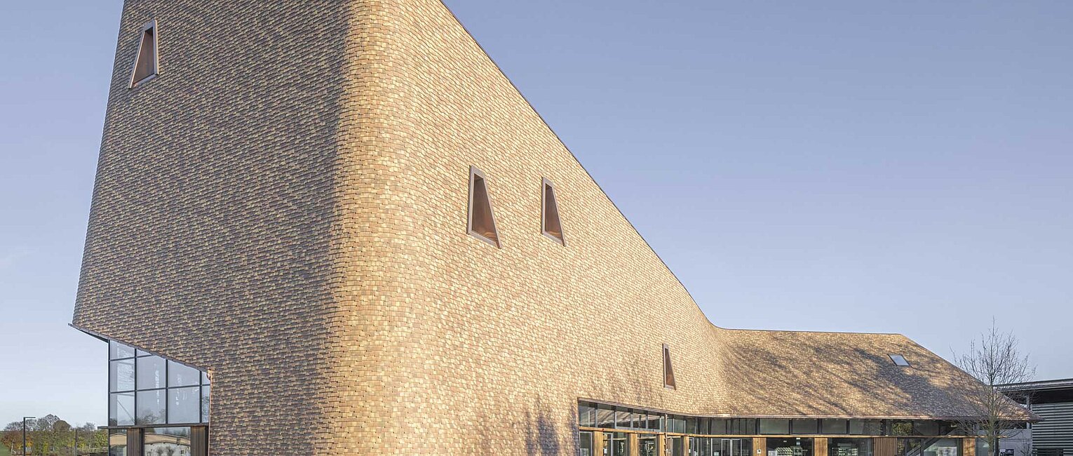 Modernes Gebäude mit geschwungener Fassade aus Holzschindeln und kleinen dreieckigen Fenstern, umgeben von Gras und offenem Himmel.