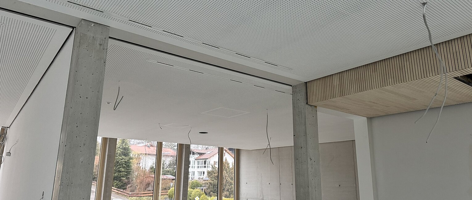 Leerer, unrenovierter Raum mit großen Fenstern, Betonpfeilern und abgehängter Decke mit sichtbaren Kabeln.