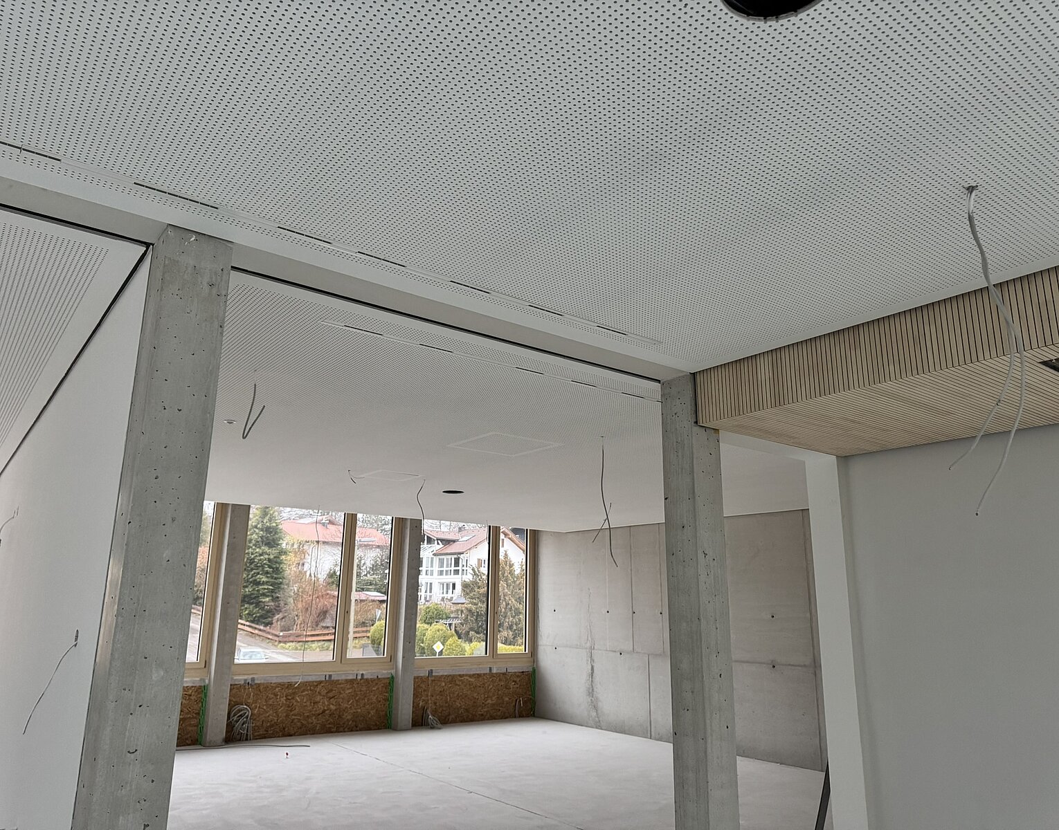 Leerer, unrenovierter Raum mit großen Fenstern, Betonpfeilern und abgehängter Decke mit sichtbaren Kabeln.